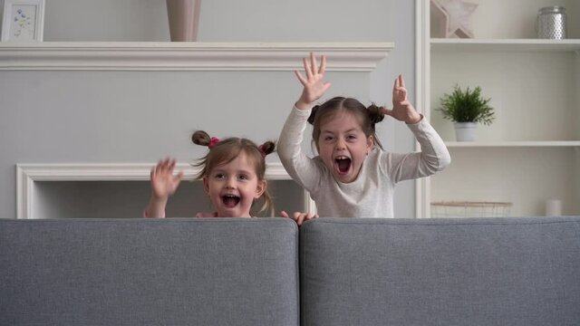 Funny little girls play at home, jump out from sofa, scare, scream and laugh, look at camera. Happy family concept.