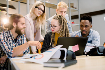 Collaboration, brainstorm. Young multiethnic people freelancers in casual wear planning business strategy. Blond girl with long dreadlocks hair is using black laptop and talking to her colleague