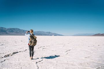 Back view of female blogger using modern smartphone for taking images while standing with touristic backpack in Badwater basin, young woman making photo of breathtaking view in Death valley lands