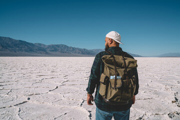 Back view of male traveler with backpack looking at wild landscape in desolate lands in death valley, hipster guy wanderlust explore nature in dry desert environment having journey on vacations.