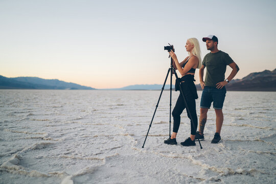 Young male and female colleagues using modern technology for taking photos of beautiful scenery of Badwater landscape, hipster girl and guy travelers shooting video on camera and tripod in journey.