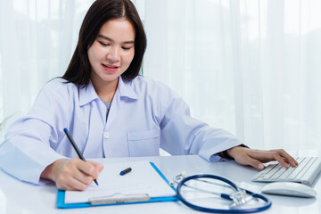 Fototapeta na wymiar Asian doctor young beautiful woman smiling using working with PC desktop computer and her writing something on paperwork or clipboard white paper at hospital desk office, Healthcare medical concept