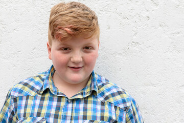 Plump red-haired boy is smiling. A boy in a plaid shirt stands on a wall background and smiles.