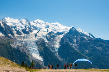 Planpraz, Chamonix, Savoie, France.  Paragliders fly, people hiking  with view of snow covered Mont Blanc mountain. Summer vacation recreation in French Alps. Active tourism background.