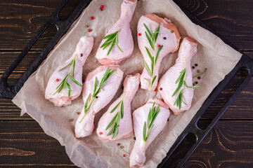 Raw chicken drumsticks on a baking sheet on parchment with rosemary and spices on a wooden background, top view