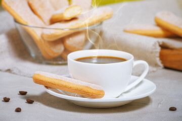 A white cup of hot aromatic coffee with steam and traditional Traditional italian savoiardi biscuits or ladyfingers cookie on a saucer and in a glass bowl. Selective focus