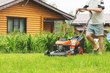 Man in shorts mowing the grass on the lawn. Young man cutting grass in his yard with lawn mower. Gardener mowing the lawn with  lawnmower in summertime - closeup.