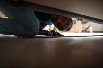 Under his bed in bedroom. African-american man looking for job in unusual places at his home. Crazy, funny way to find career and going up. Concept of crisis, unemployment, finance, business.