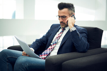 Fototapeta premium Confident male CEO having phone conversation with financier checking accounting quarter report discussing sum of spending costs while sitting in light cabinet with free wireless connection to internet