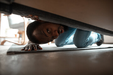 Under his bed in bedroom. African-american man looking for job in unusual places at his home....