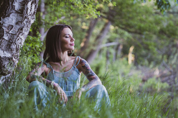 beautiful natural woman with tattoos sitting on the grass, leaning on her knees on the shore of a forest lake, wearing a blue dress and looking at the water