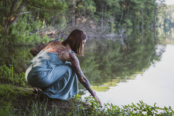 beautiful natural woman with tattoos squatting sideways on the grass on the shore of a forest lake, wearing a blue dress and reaching into the water