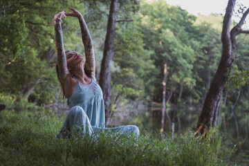 beautiful natural woman with tattoos sitting on the grass on the shores of a forest lake, stalling - obrazy, fototapety, plakaty