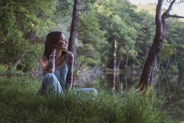 beautiful natural woman with tattoos sitting on the grass on the shore of a forest lake
