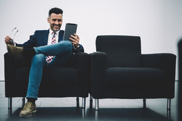 Cheerful successful proud ceo dressed in formal wear watching funny videos on touch pad connected to 4G internet sitting on armchair.Positive mature entrepreneur laughing while reading news on tablet