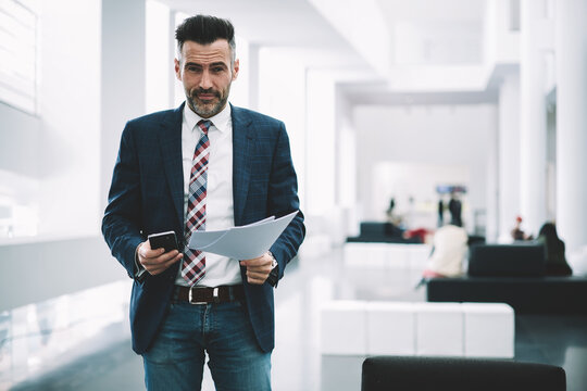 Portrait of middle aged successful proud ceo dressed in formal wear smiling at camera while holding financial documents in hands.Prosperous executive director with smartphone standing in office