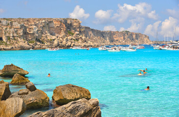 paradise clear torquoise blue water with boats and cloudy blue sky in background in Favignana island, Cala Rossa Beach, Sicily South Italy.