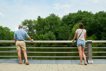 A bald man and a woman enjoying the view of the river keeping distance a few meters to avoid the spread of coronavirus. A guy and girl in face masks resting leaning against the fence on the promenade