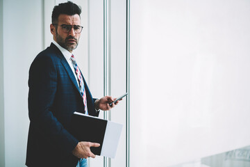 Half length portrait of confident mature entrepreneur dressed in formal wear checking mail on smartphone device connected to 4G internet standing near publicity area for your business information