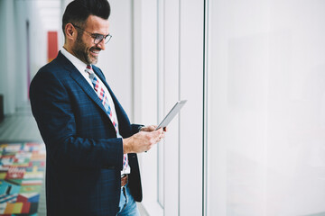 Cheerful handsome executive manager watching funny videos on touchpad having work  break standing near advertising area, positive CEO chatting with employees using wireless connection in office