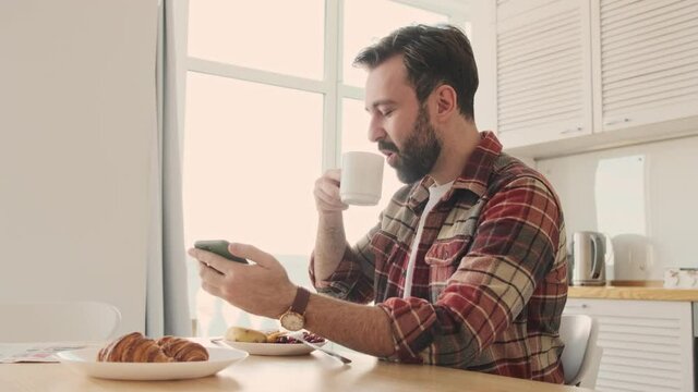 Handsome young positive optimistic bearded man at the kitchen indoors at home having a breakfast while using cellphone