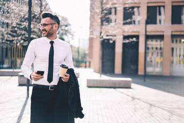 Smiling bearded businessman dressed in formal wear sending text message to colleague using wireless internet connection walking near urban building with coffee, copy space for your advertise