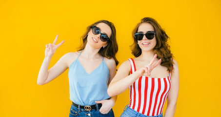 Two young beautiful blond smiling hipster girls in trendy summer clothes. Sexy carefree women posing near yellow wall in sunglasses. Positive models going crazy