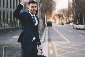 Handsome mature bearded manager of trading corporation hailing taxi raising hand standing on road in New York city street near copy space area for your advertising getting to business meeting