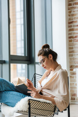 Image of focused woman reading book while sitting in armchair
