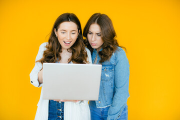 Portrait of two shocked young blackhead girls pointing finger at laptop computer isolated over yellow background