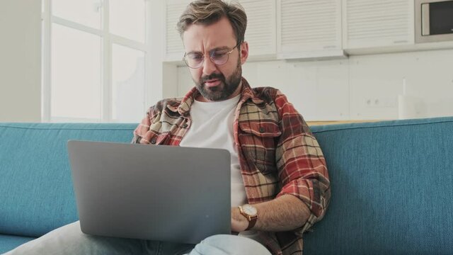 Handsome young tired bearded man indoors at home sitting on a sofa while using laptop computer
