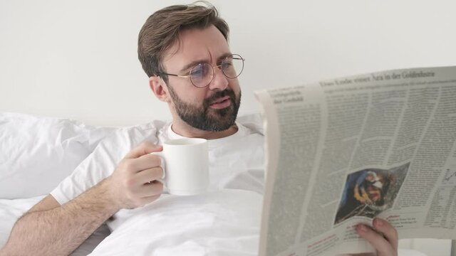 Handsome young concentrated bearded man indoors at home lies in bed while reading newspaper and drinking coffee