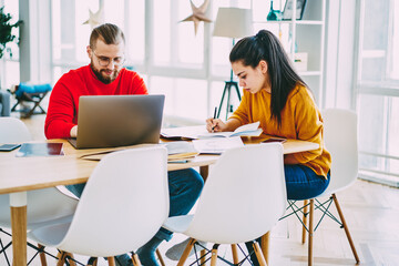 Young male and female colleagues concentrated on working process making research online and writing, students spending time in coworking space together , man using laptop while woman make notes.