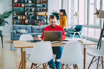 Smiling hipster guy enjoying distance job sitting at desktop at home interior while his wife talking on mobile phone, cheerful young man satisfied with freelance work blogging via laptop computer.
