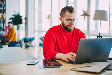 Skilled male freelancer working at home interior using laptop computer and wireless internet sitting at desktop, hipster guy watching online video on netbook, man checking mail and blogging .