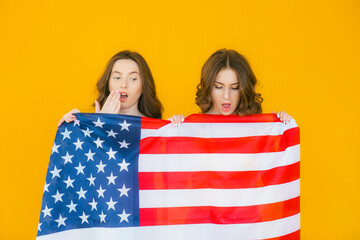 American girls. Two playful young women gesturing peace sign and holding American flag while standing on yellow background