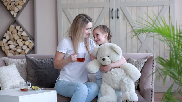 Happy mother and daughter sitting at home on the couch in white t-shirts. A woman drinks tea.A child hugs a large Teddy bear. The mother gently embraces the child