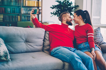 Young marriage posing for picture on smartphone camera looking at each other smiling and hugging at living room, positive couple in love taking romantic pictures feeling happiness of being together.