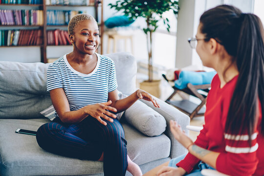 Cheerful african american woman talking with female friend spending time together at apartment, multiracial hipster girls having conversation at home interior, female explaining emotional gesture .