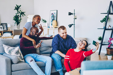 Naklejka premium Positive multiracial friends having fun at apartment posing for selfie on smartphone camera, young cheerful male and female hipsters taking photo on mobile phone during meeting at home interior.