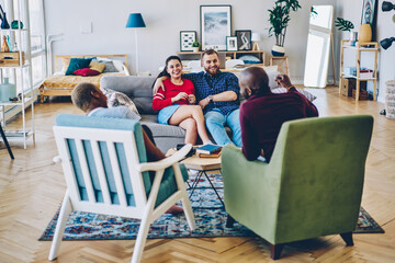 Cheerful romantic couple sitting at couch in living room having conversation and being hospitable to guests, four multiracial friends communicating and having fun on meeting at home interior.