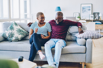Obraz premium Happy african american couple laughing at funny joke sitting on couch in living room, cheerful man feeling carefree tith positive wife resting on weekend together in modern designed apartment.