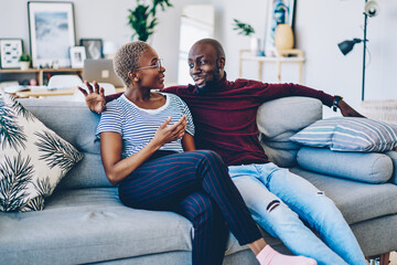 Obraz premium Positive african american couple in love sitting on comfortable couch having conversation about relationships,young dark skinned family resting at home together communicating at living room .