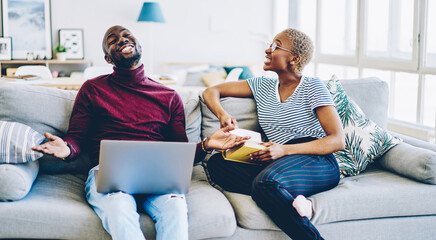 Happy young marriage having conversation on laughing while spending time on hobby during weekend with book and laptop computer, cheerful african american couple in love having fun together.