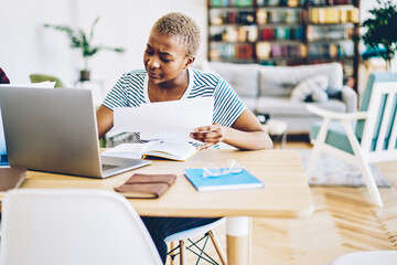 Obraz premium Concentrated african american woman making banking online using papers w with information, serious skilled dark skinned female freelancer doing remote job on laptop computer at home interior