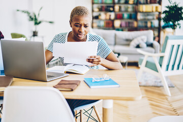 Sad african american woman reading document having trouble with bills for wifi, upset hipster girl preparing for exams in university learning information sitting at desktop in stylish home interior.