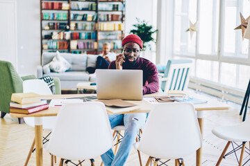 Serious dark skinned hipster guy talking on mobile phone while confirming booking on website, skilled african american freelancer concentrated on online business making telephone call and browsing.