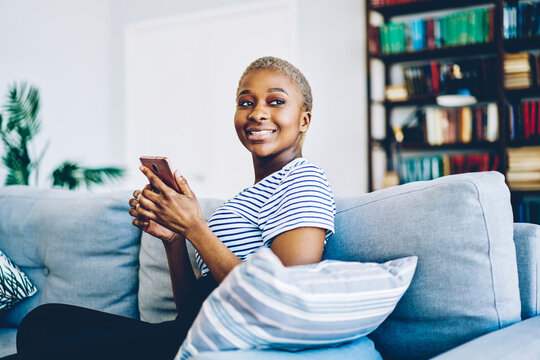 Cheerful african american woman satisfied with wireless internet at home connecting smartphone for chatting and blogging,happy dark skinned hipster girl using mobile phone for sending message.