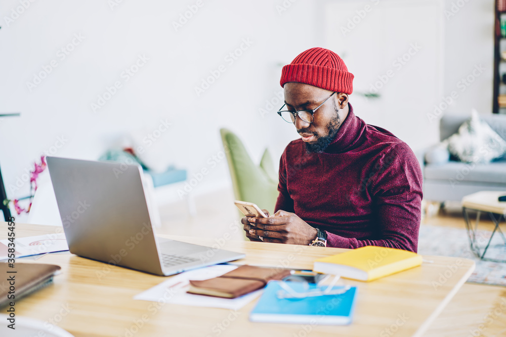 Poster serious african american hipster guy sending text message via smartphone while sitting at desktop wi