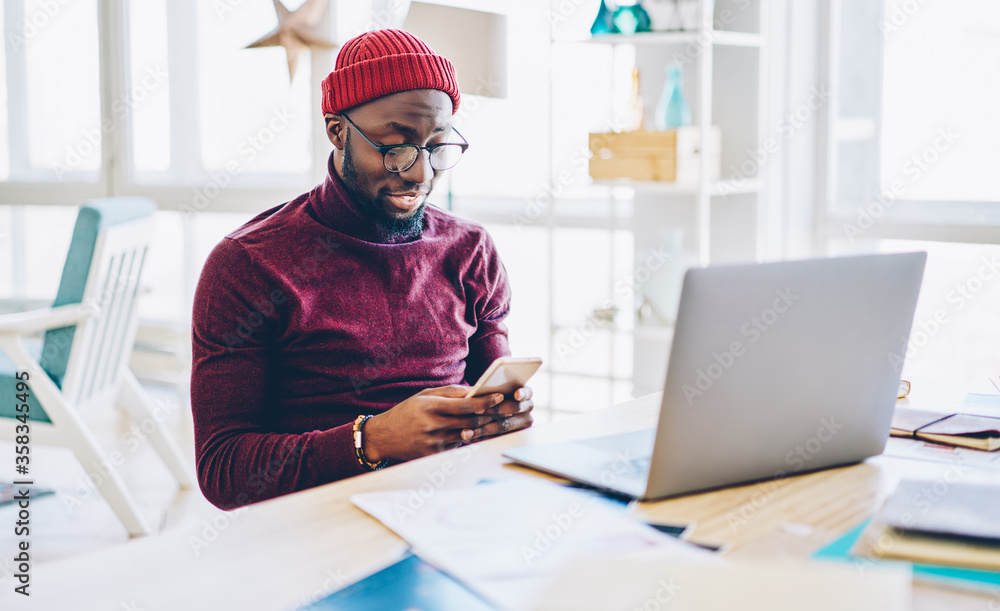Poster african american hipster guy in spectacles sending text message on mobile phone while working online